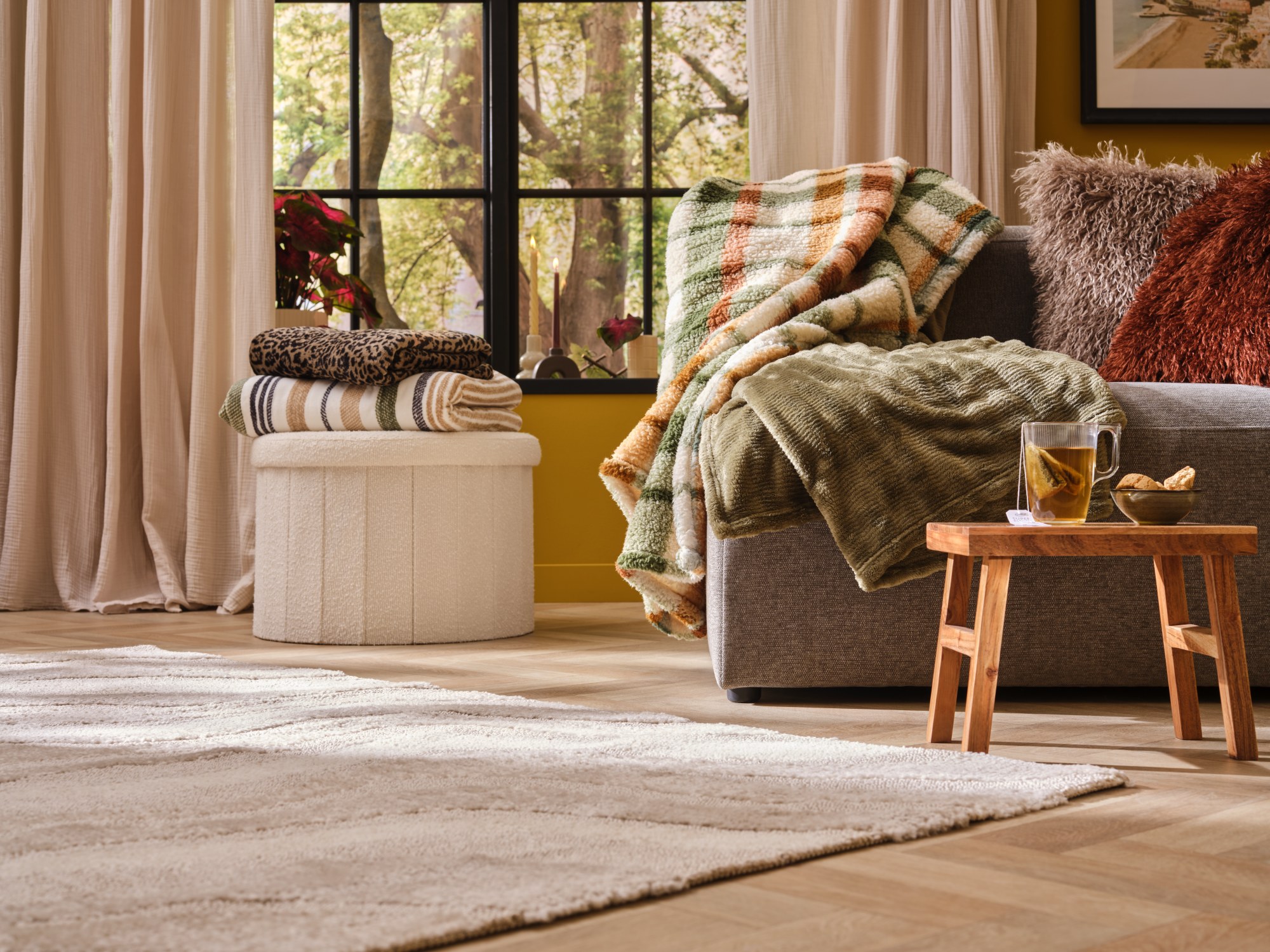 Plaid a quadri e coperta in pile verde drappeggiati sul bracciolo di un divano grigio, con plaid ripiegati su un pouf