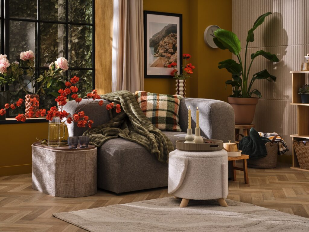 Soggiorno autunnale con divano grigio, plaid verde, cuscino a quadri, pouf in bouclé e tappeto su parquet a spina di pesce