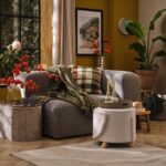 Soggiorno autunnale con divano grigio, plaid verde, cuscino a quadri, pouf in bouclé e tappeto su parquet a spina di pesce
