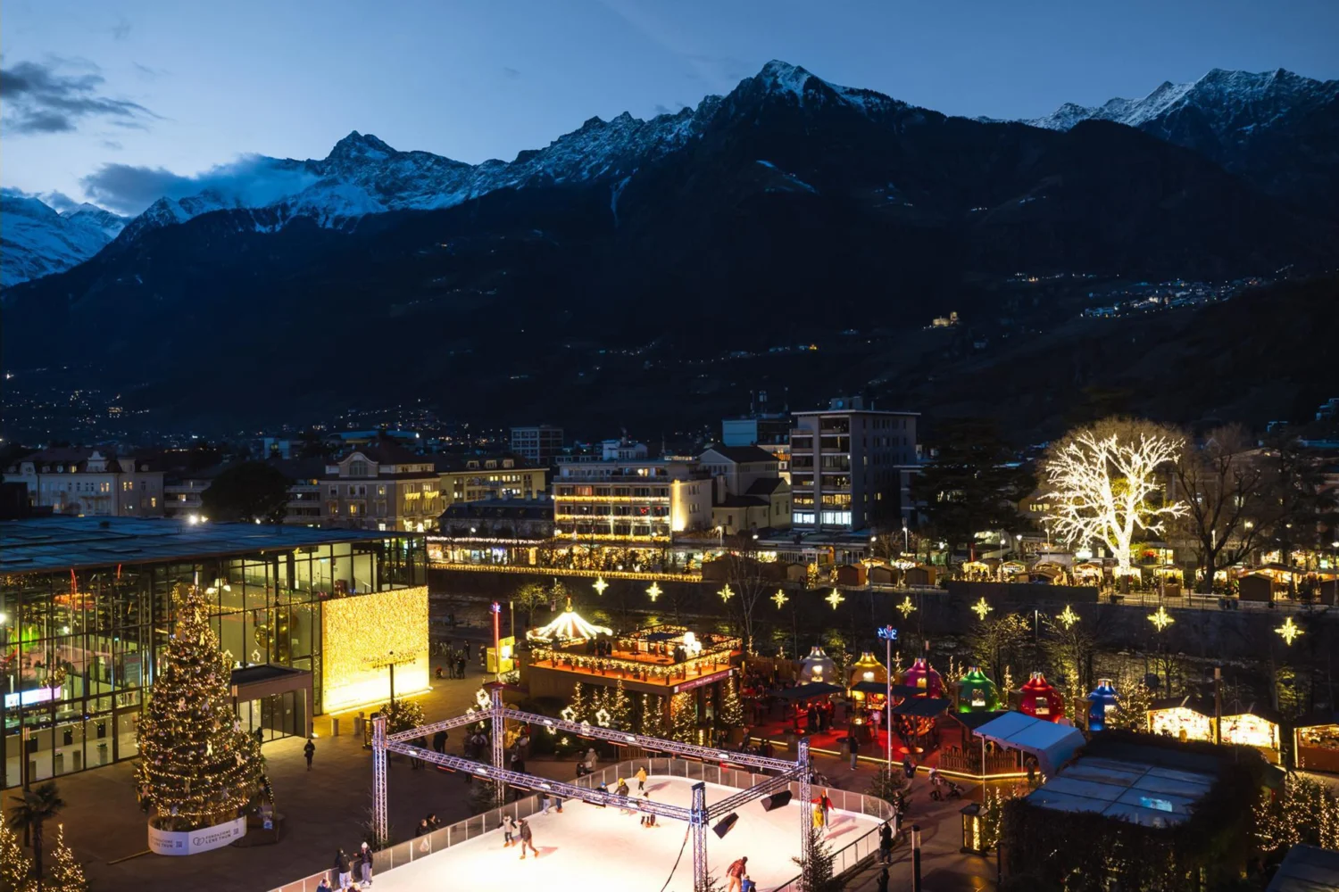 Piazza Terme in Merano at Christmas with the ice rink and the mountains at dusk